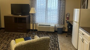 Habitación de hotel con televisor de pantalla plana sobre un mueble de madera, alfombra estampada, silla de mimbre con cojines, lámpara de pie, ventana con cortinas estampadas, unidad de aire acondicionado, refrigerador y extintor de incendios en la pared.