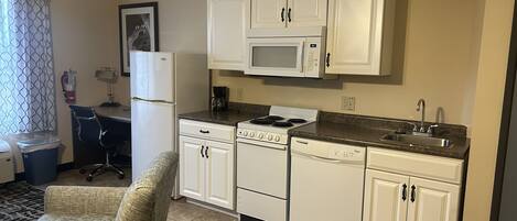 A compact kitchen with white cabinets, a microwave, a stove, and a sink. There is a chair and a small rug in the seating area.