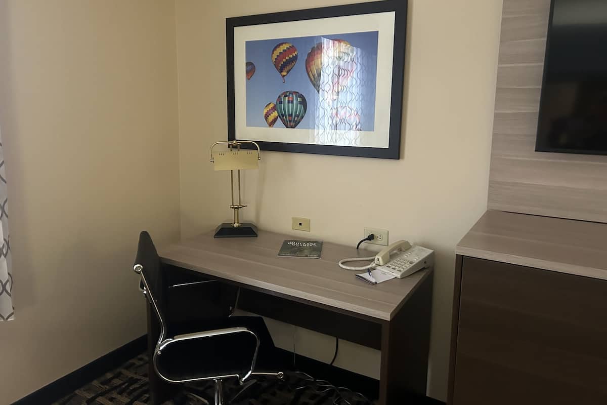 A hotel room with a desk, a chair, a framed picture of hot air balloons, a lamp, a telephone, and a television.