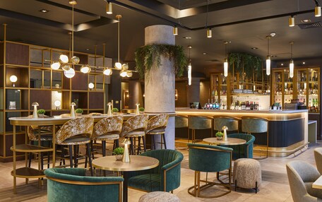 A modern bar area with round tables, green upholstered chairs, and a bar counter with a variety of bottles.
