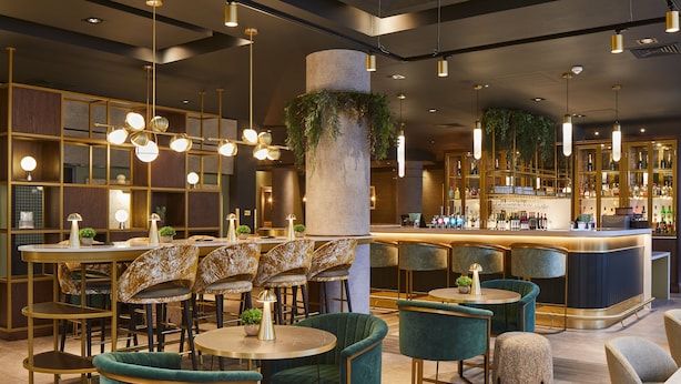 A modern bar area with round tables, green upholstered chairs, and a bar counter with a variety of bottles. - Leonardo Royal Hotel Glasgow (Glasgow)