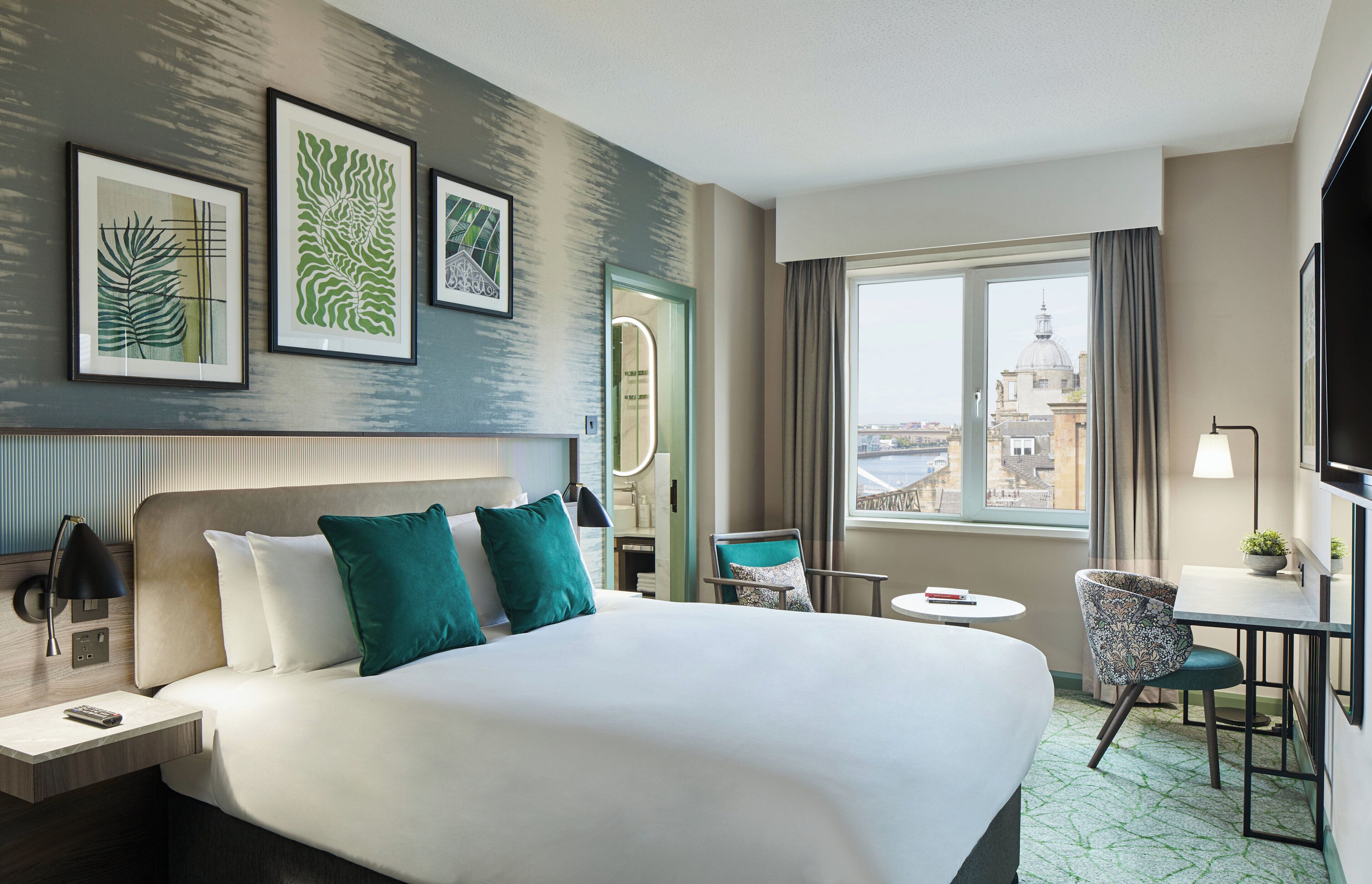 A hotel room with a large bed, a desk, a chair, and a view of the city.