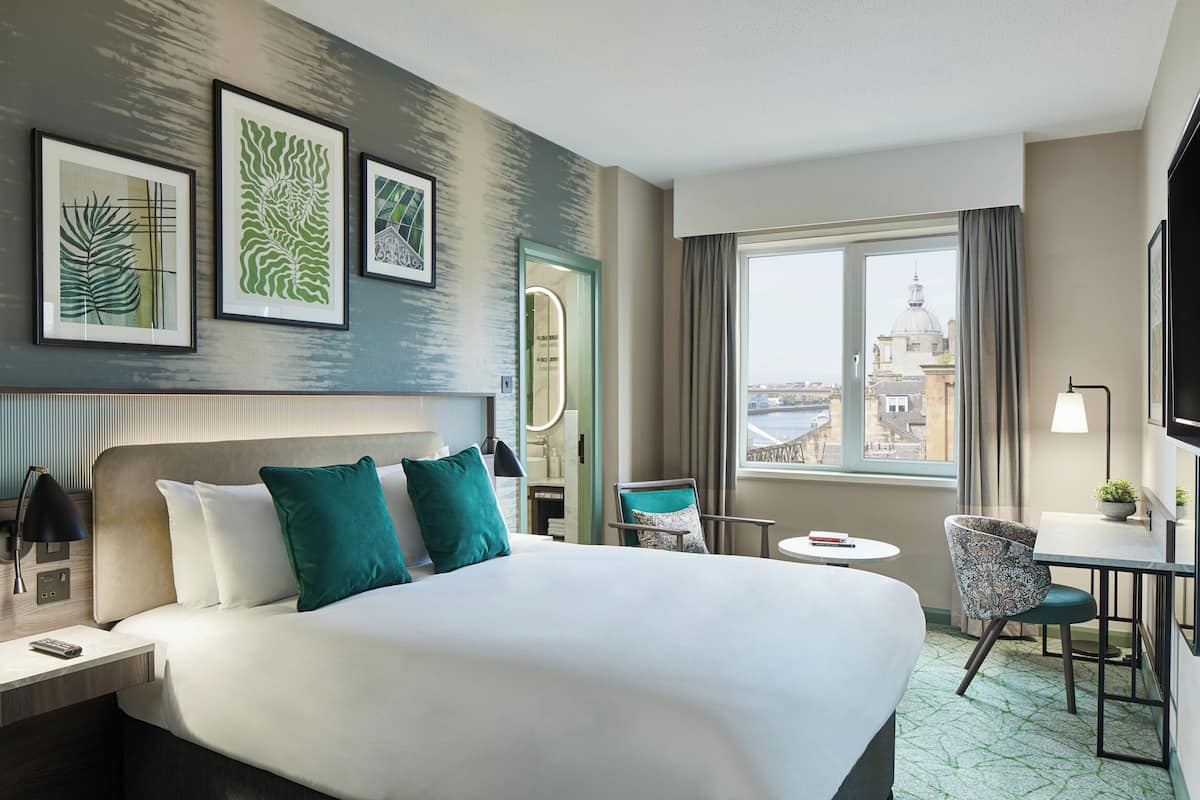 A hotel room with a large bed, a desk with a chair, a small table, and a window with a view of a cityscape.