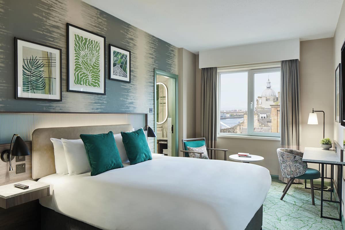 A hotel room with a large bed, a desk with a chair, a small table, and a window with a view of a cityscape.