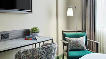Habitación de hotel con una cama, un escritorio con una planta, una silla con cojín estampado, una lámpara de pie y un televisor en la pared.
