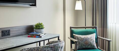 A hotel room with a bed, a desk with a plant, a chair with a patterned cushion, a standing lamp, and a television on the wall.