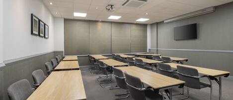 A conference room with multiple tables and chairs, a projector, a flat-screen TV, and framed pictures on the wall.
