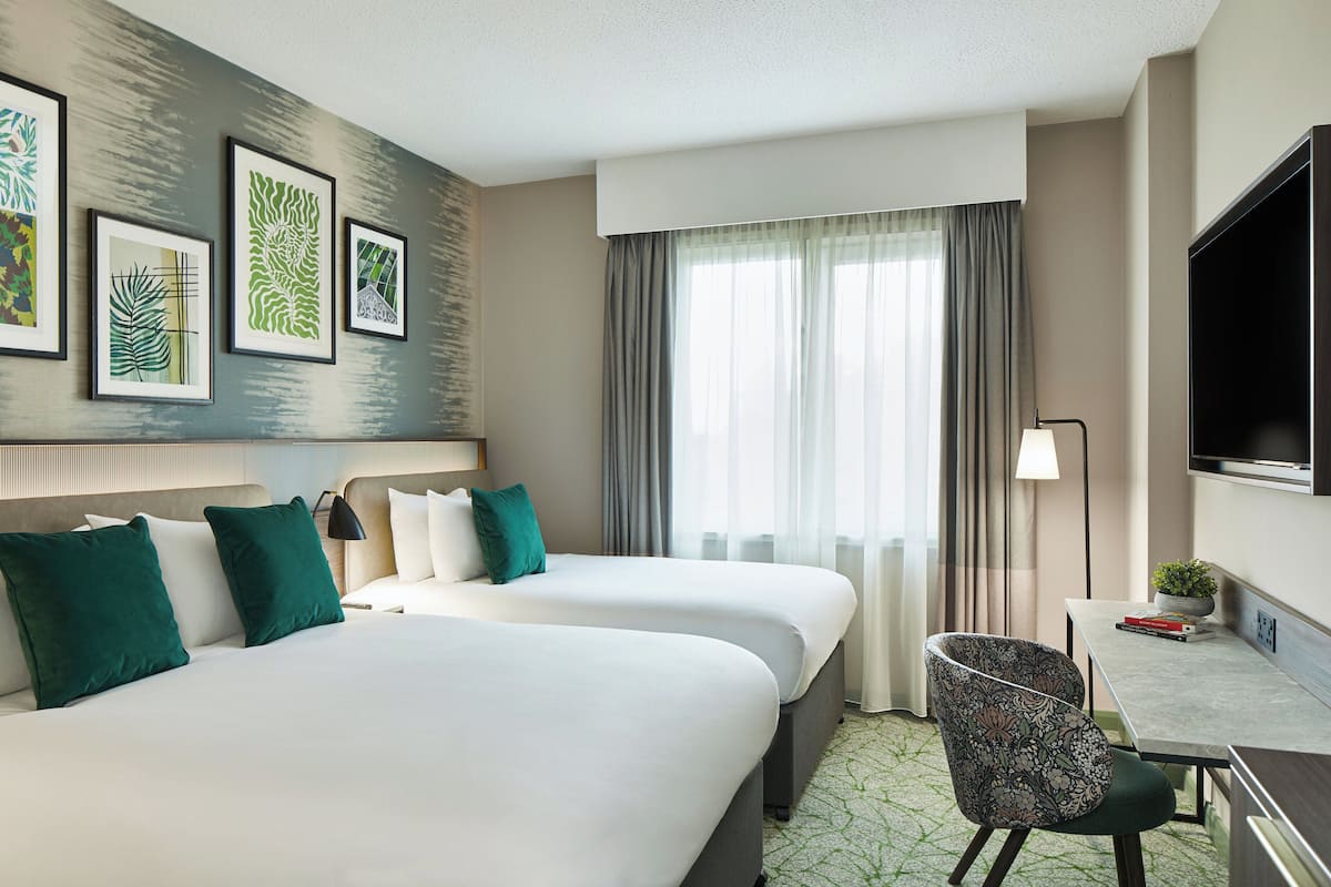 A hotel room with two beds, a desk with a chair, a TV, and artwork on the wall.