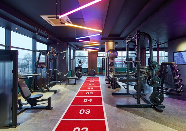 A gym with various exercise equipment, a red running track, and large windows offering a city view.