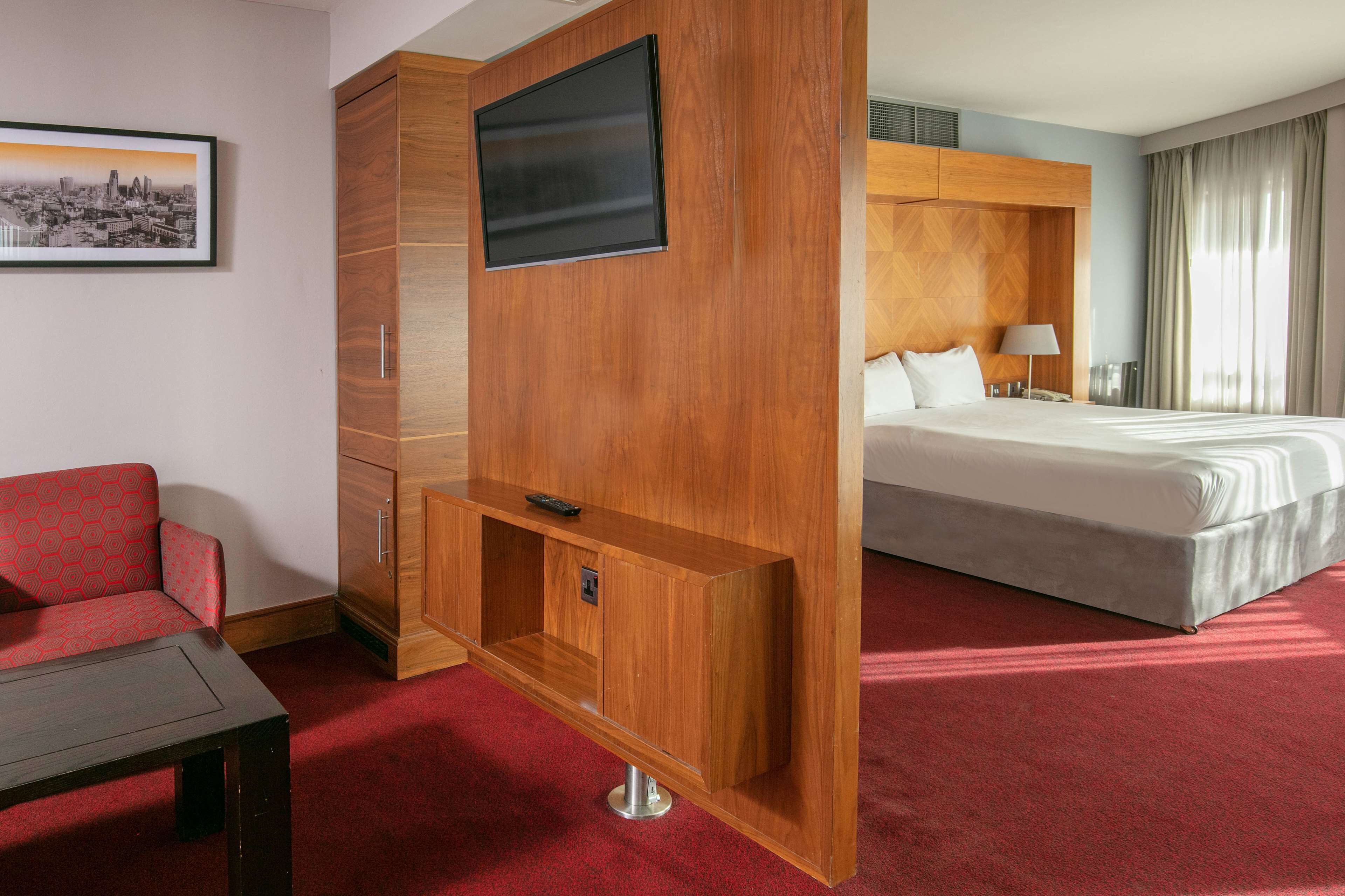 A hotel room with a bed, a TV mounted on a wooden panel, a red sofa, and a small table.