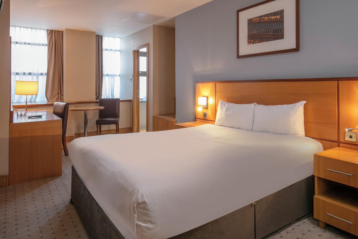 A hotel room with a large bed, a desk with a lamp, a chair, a bedside table with a lamp, and a framed picture on the wall.