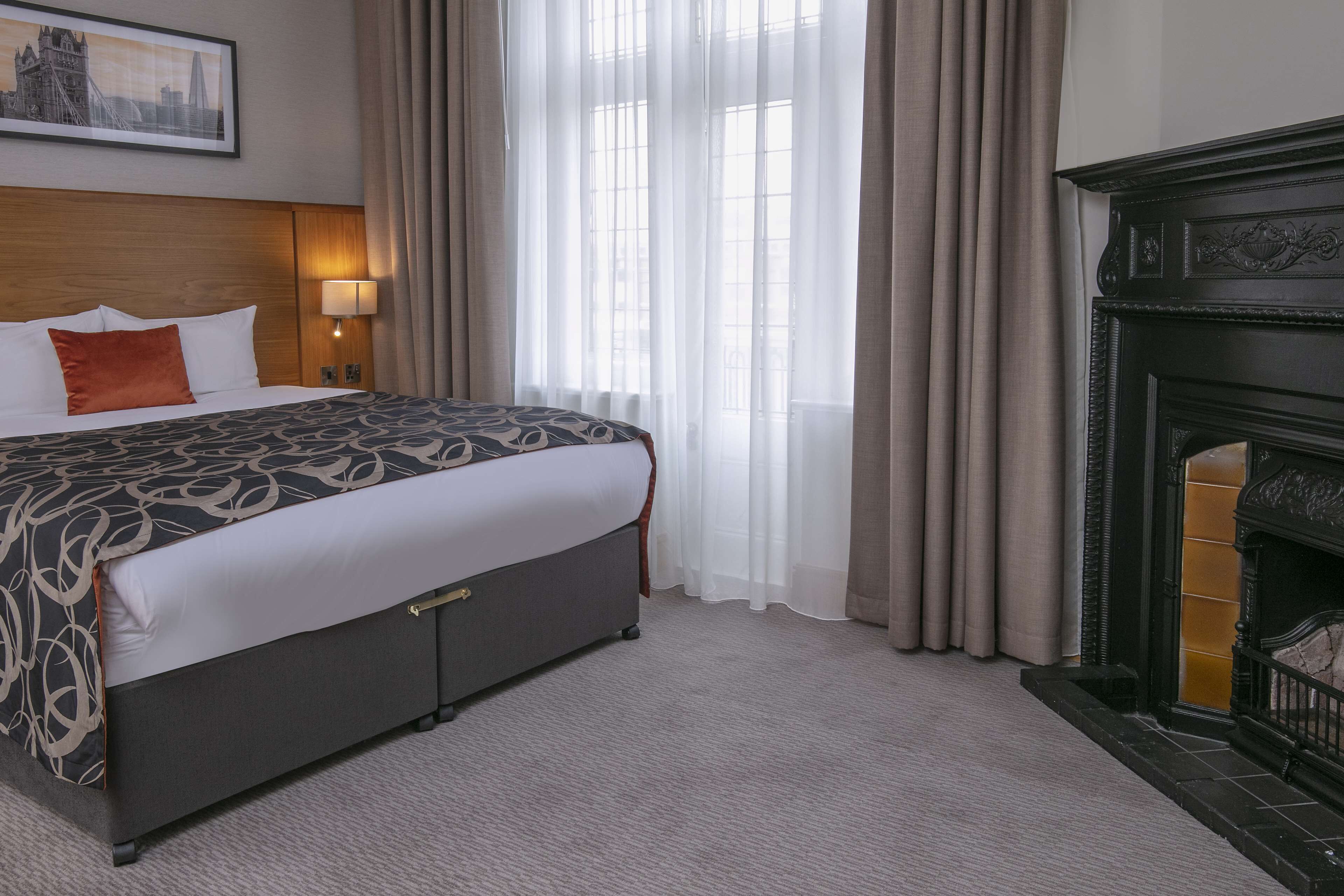 Habitación de hotel con una cama grande, una mesita de noche con lámpara, chimenea y ventana con cortinas traslúcidas.