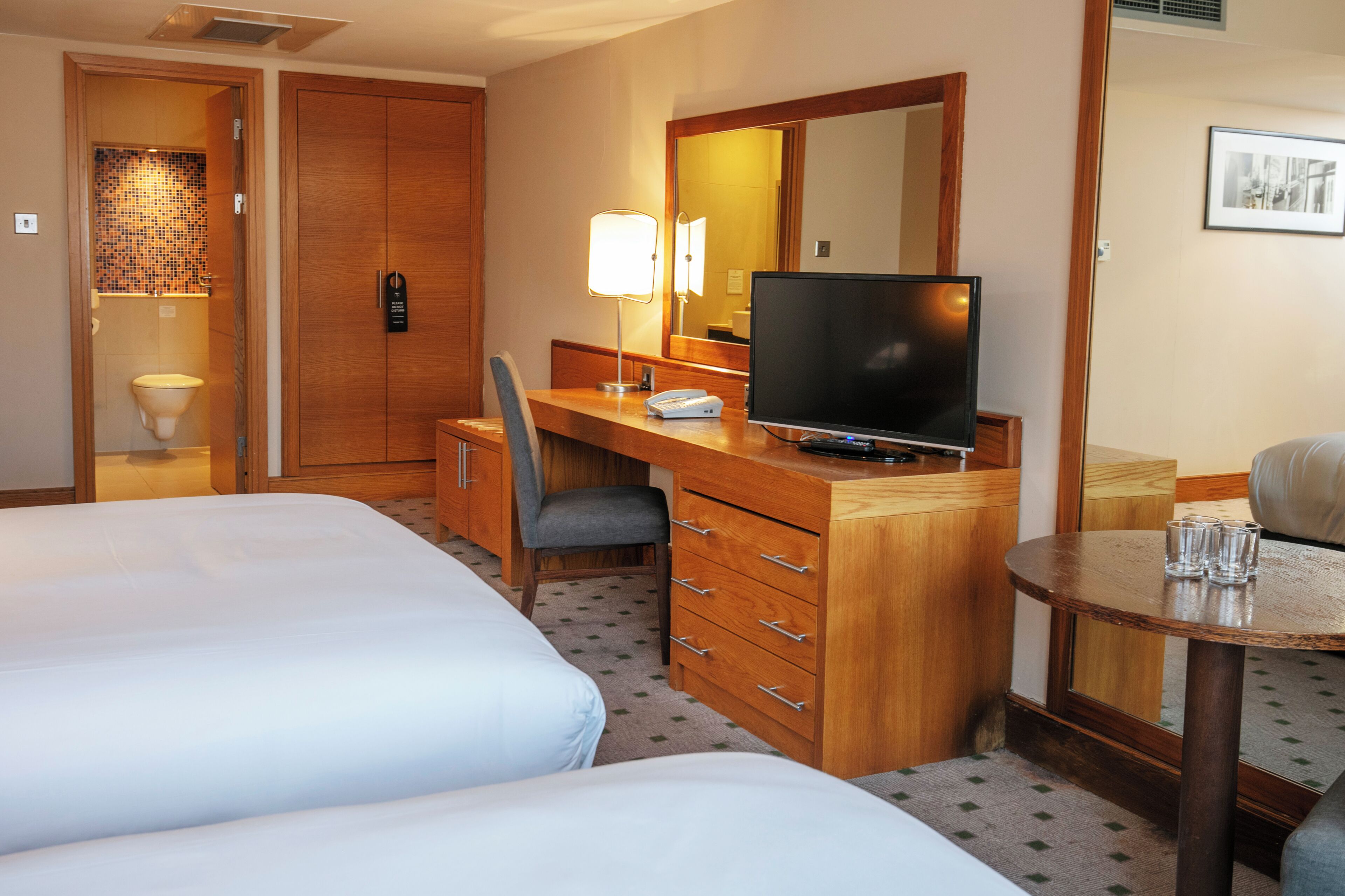 A hotel room with two beds, a desk with a television, a chair, a small table with glasses, and a mirror.