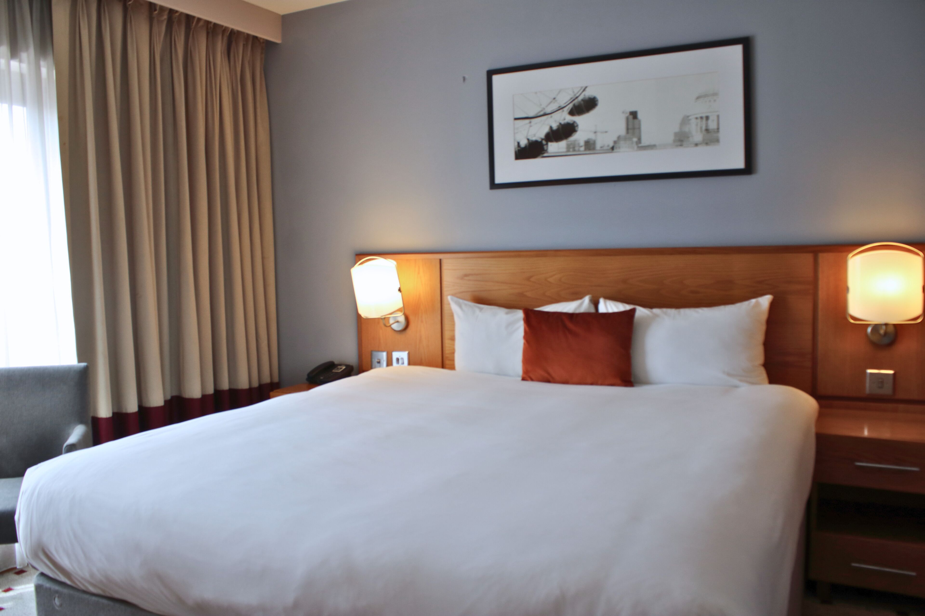 Ein Hotelzimmer mit einem großen Bett, zwei Nachttischlampen, einem gerahmten Bild an der Wand und einem Fenster mit Vorhängen.