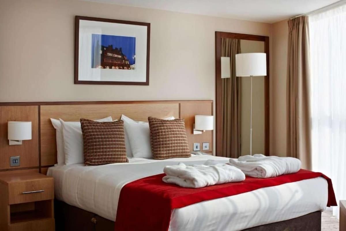 Een hotelkamer met een bed, kussens, een rode deken, een nachtkastje, een lamp, een spiegel en een fotokader aan de muur.