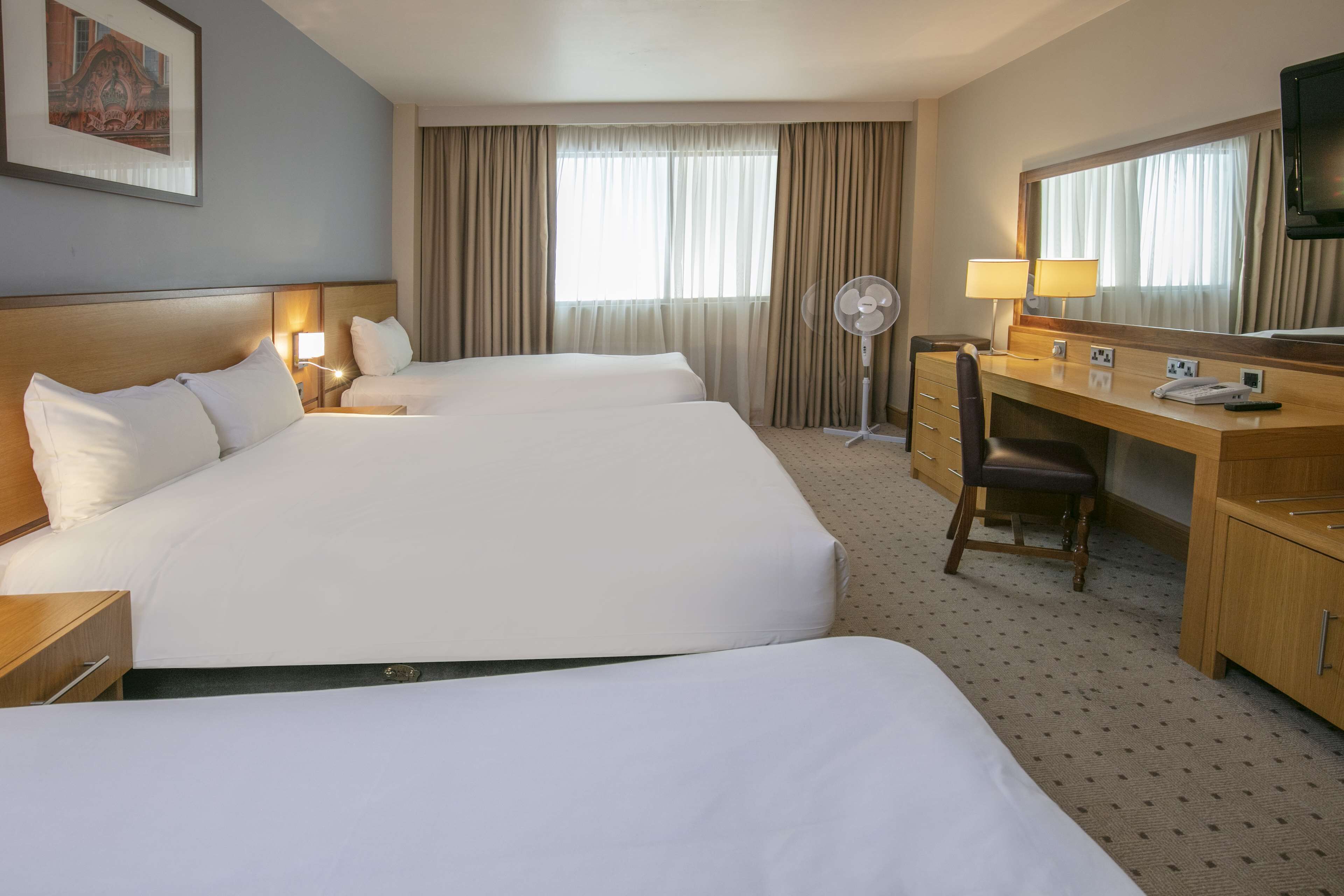 A hotel room with two large beds, a desk with a chair, a desk lamp, a flat-screen TV, and a large mirror.