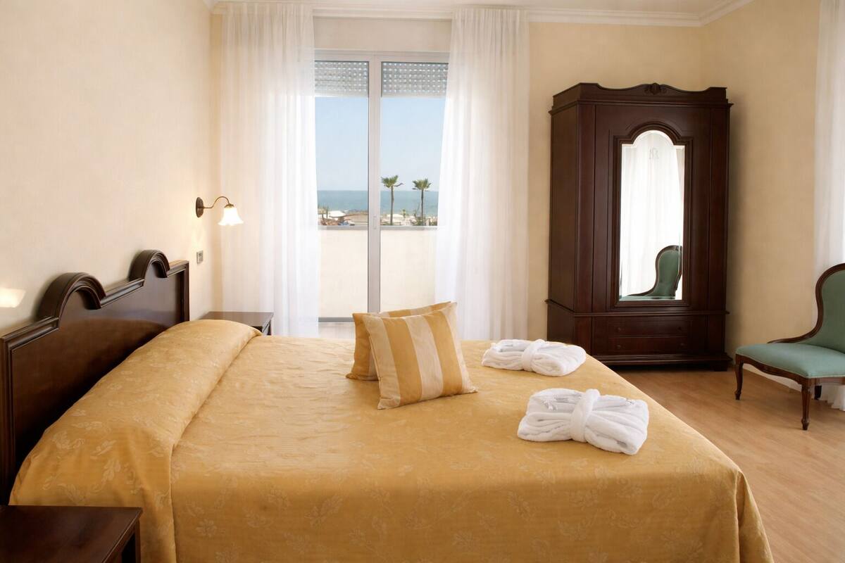 A hotel room with a bed, a large mirror, a green chair, and a view of the sea through the window.