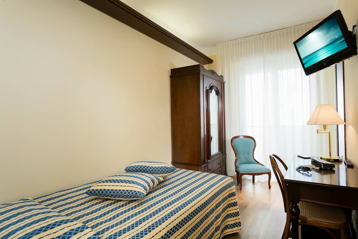 A hotel room with a bed, a desk with a lamp, a chair, a television mounted on the wall, and a wardrobe.