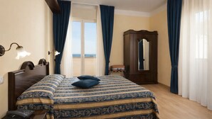 A hotel room with a bed, a wooden wardrobe, a bedside telephone, and a view of the sea through a large window.