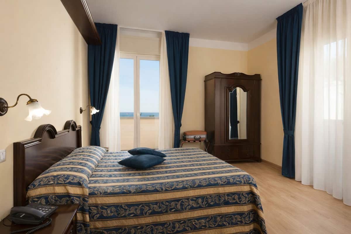 A hotel room with a bed, a wooden wardrobe, a bedside telephone, and a view of the sea through a large window.