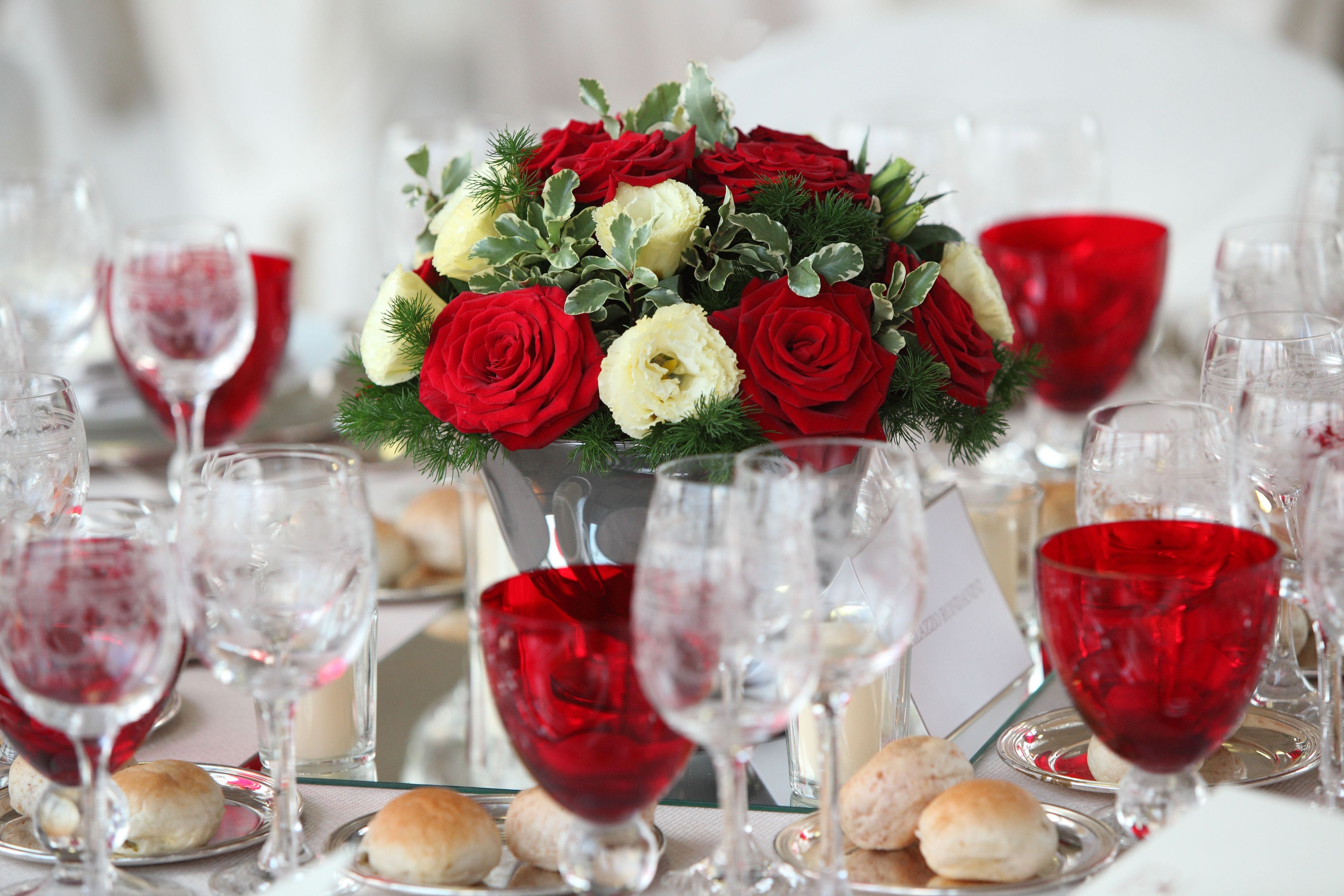 A table set with red wine glasses, a bouquet of red and white roses, and small pastries.