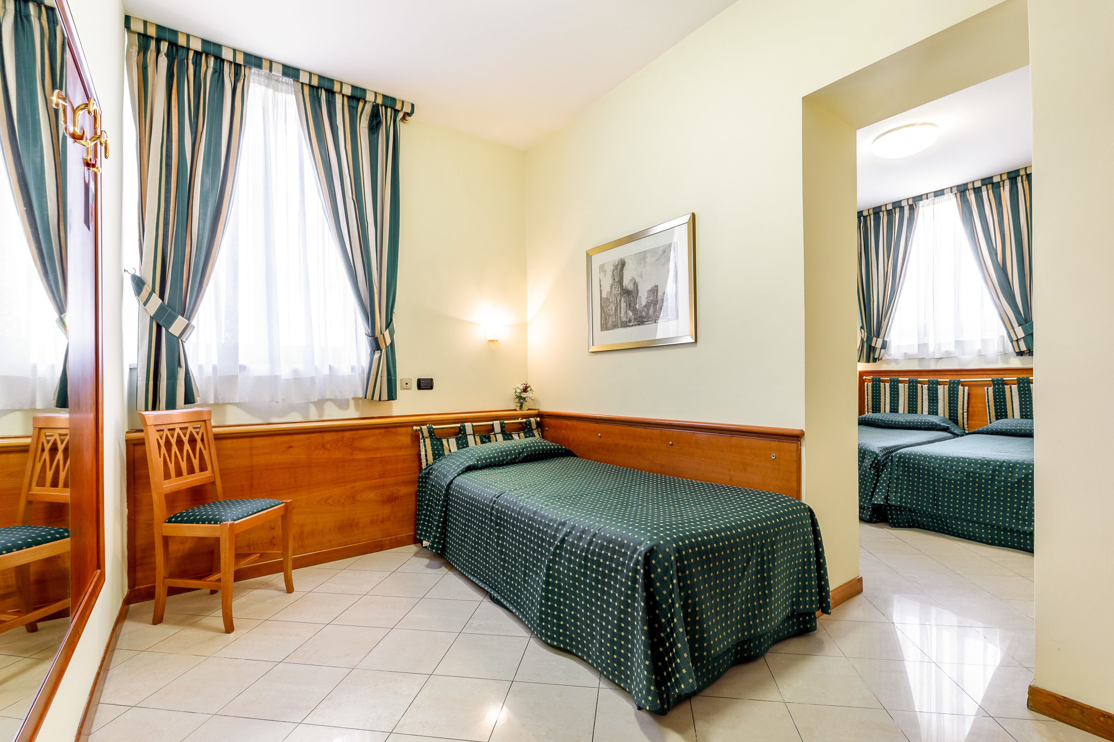 Une chambre d’hôtel avec un lit, une chaise, une table de chevet, un miroir et une fenêtre avec des rideaux.