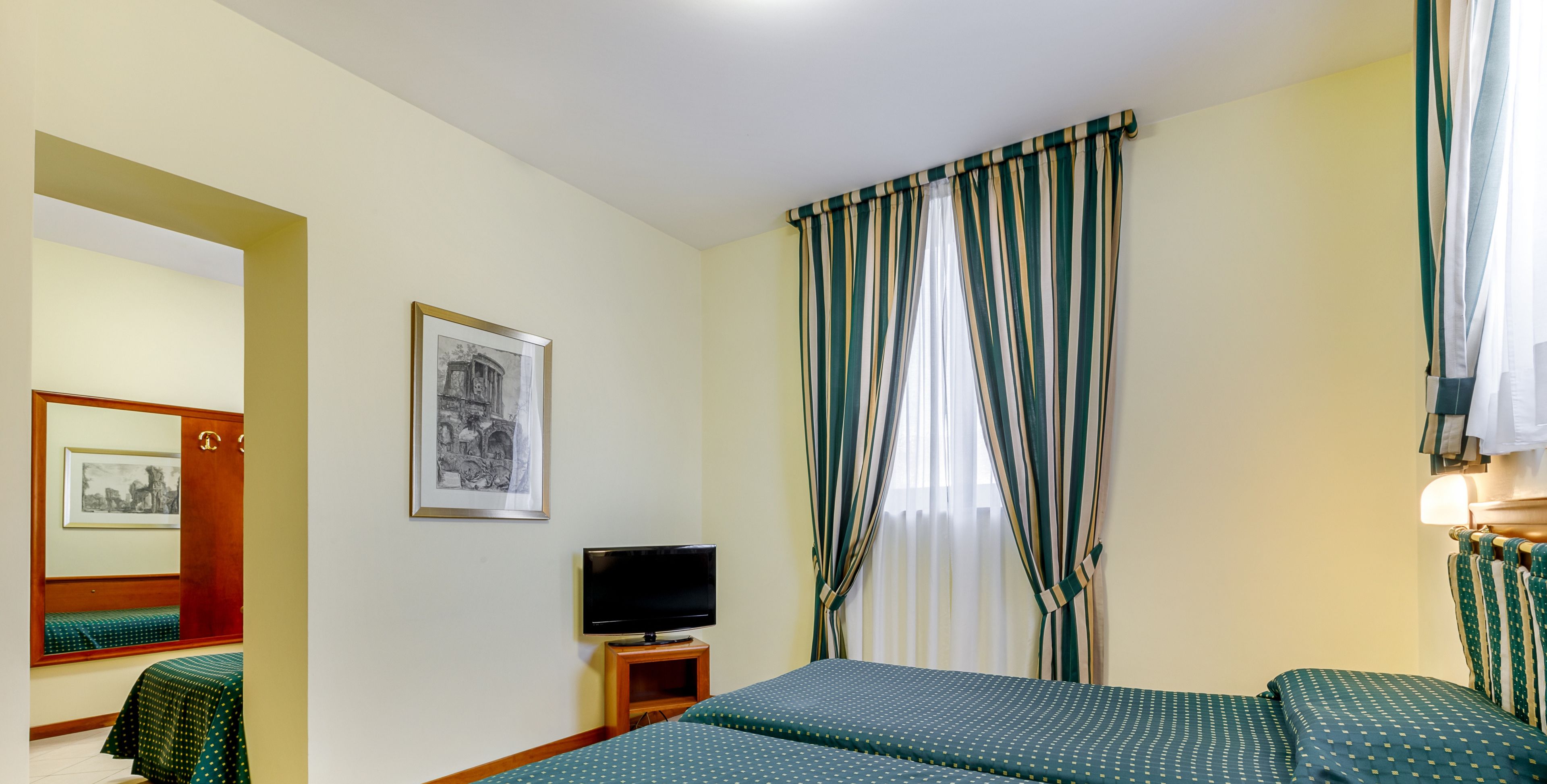 Habitació d'hotel amb un llit, un televisor sobre un suport, un quadre amb marc a la paret i una finestra amb cortines.