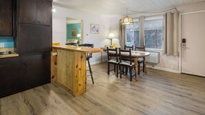 Une cuisine avec des meubles de rangement en bois, un îlot central en bois, une table à manger avec des chaises, un luminaire suspendu et une fenêtre avec des rideaux.