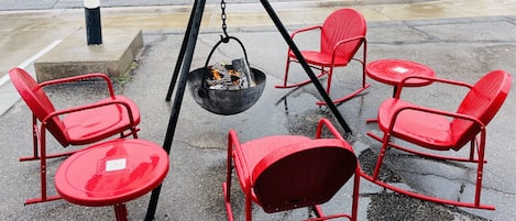 A set of red metal chairs and tables arranged on a wet pavement, with a black metal bowl containing a fire on a tripod stand.