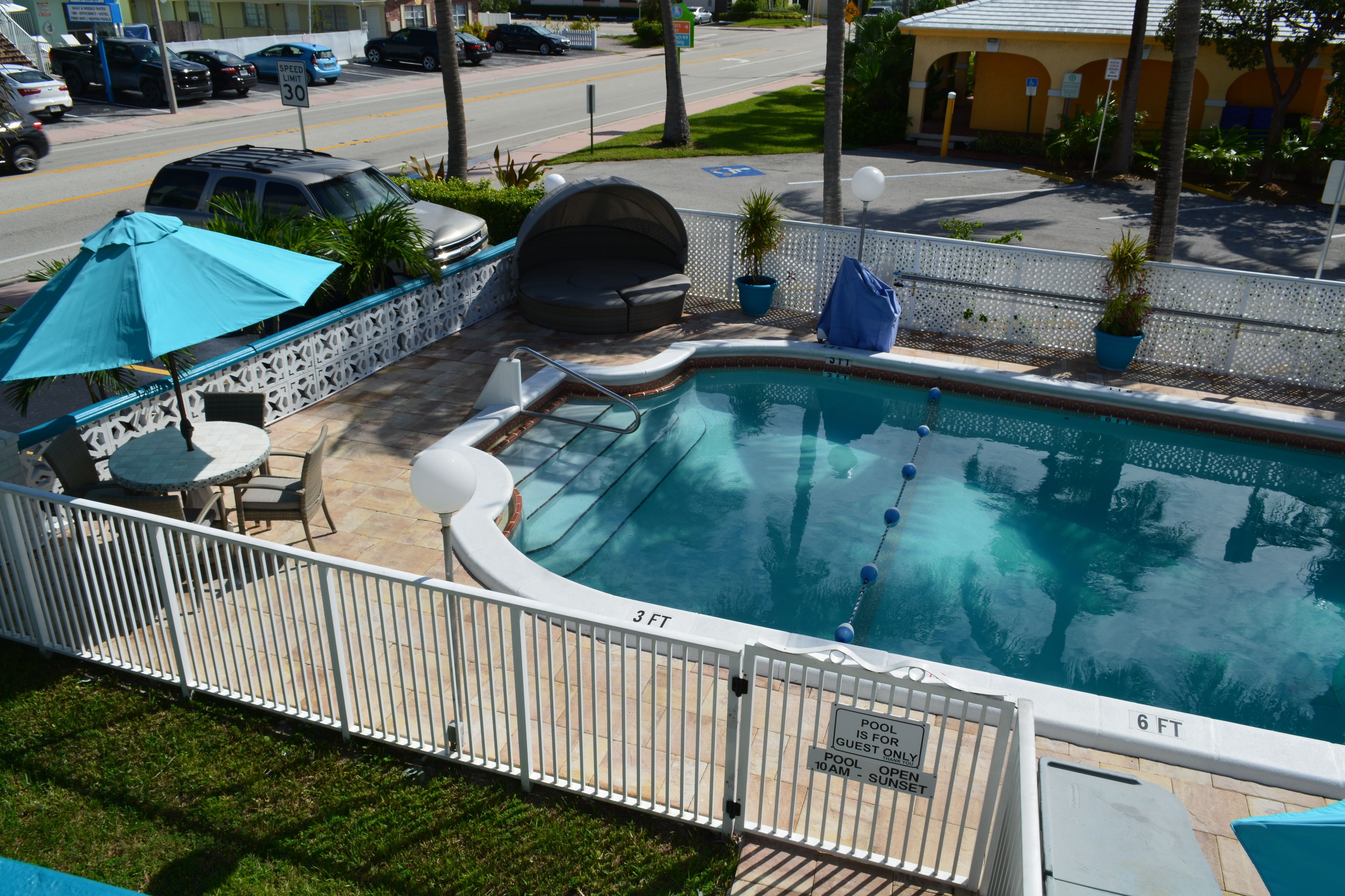 A swimming pool with a 3 FT and 6 FT depth marking, surrounded by a white fence, with a patio area including chairs and umbrellas.