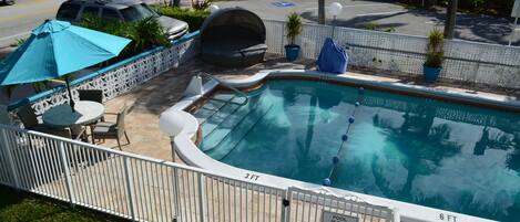 A swimming pool with a 3 FT and 6 FT depth marking, surrounded by a white fence, with a patio area including chairs and umbrellas.
