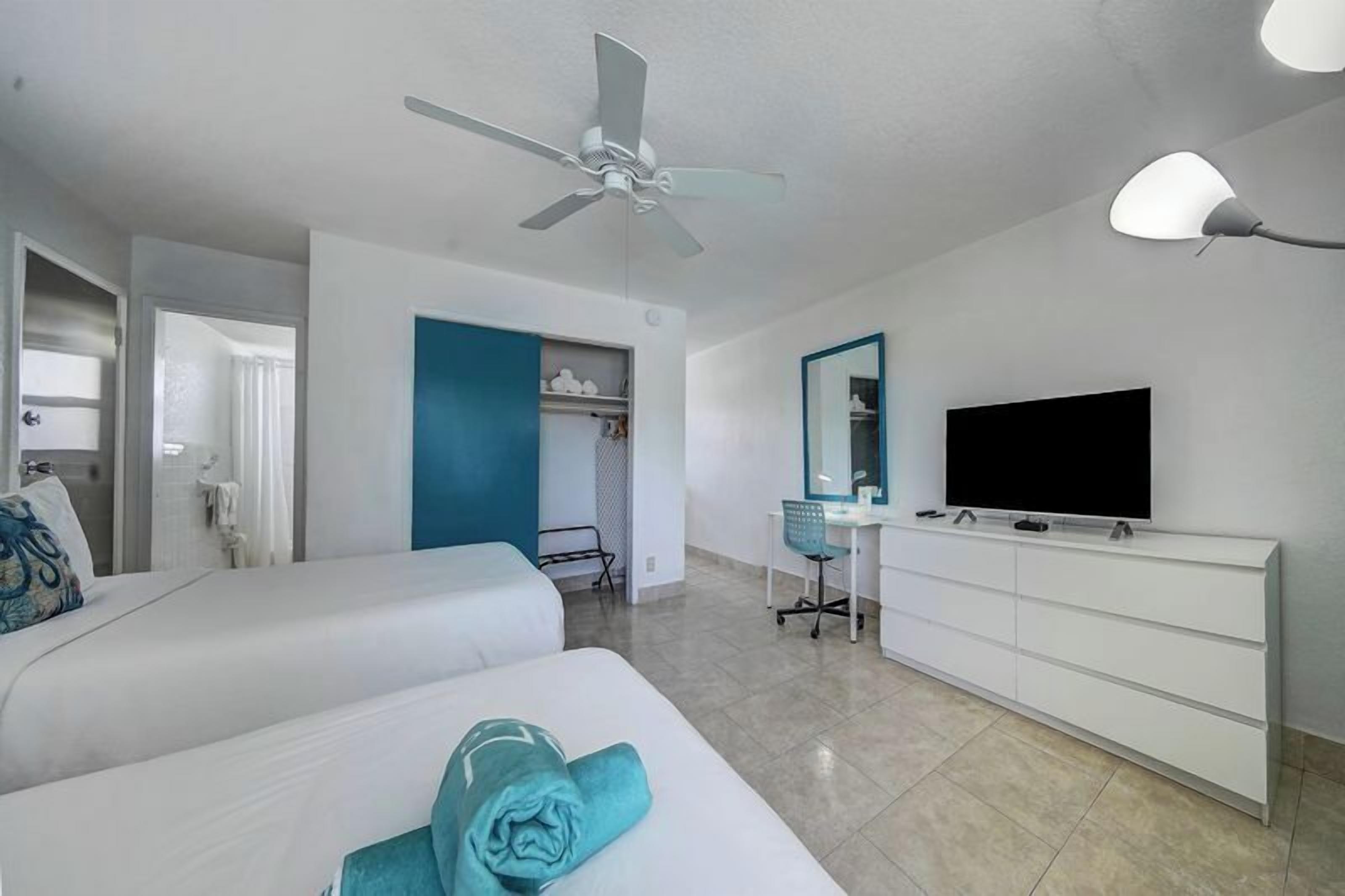 A hotel room with two beds, a desk, a TV, a ceiling fan, and a wardrobe.
