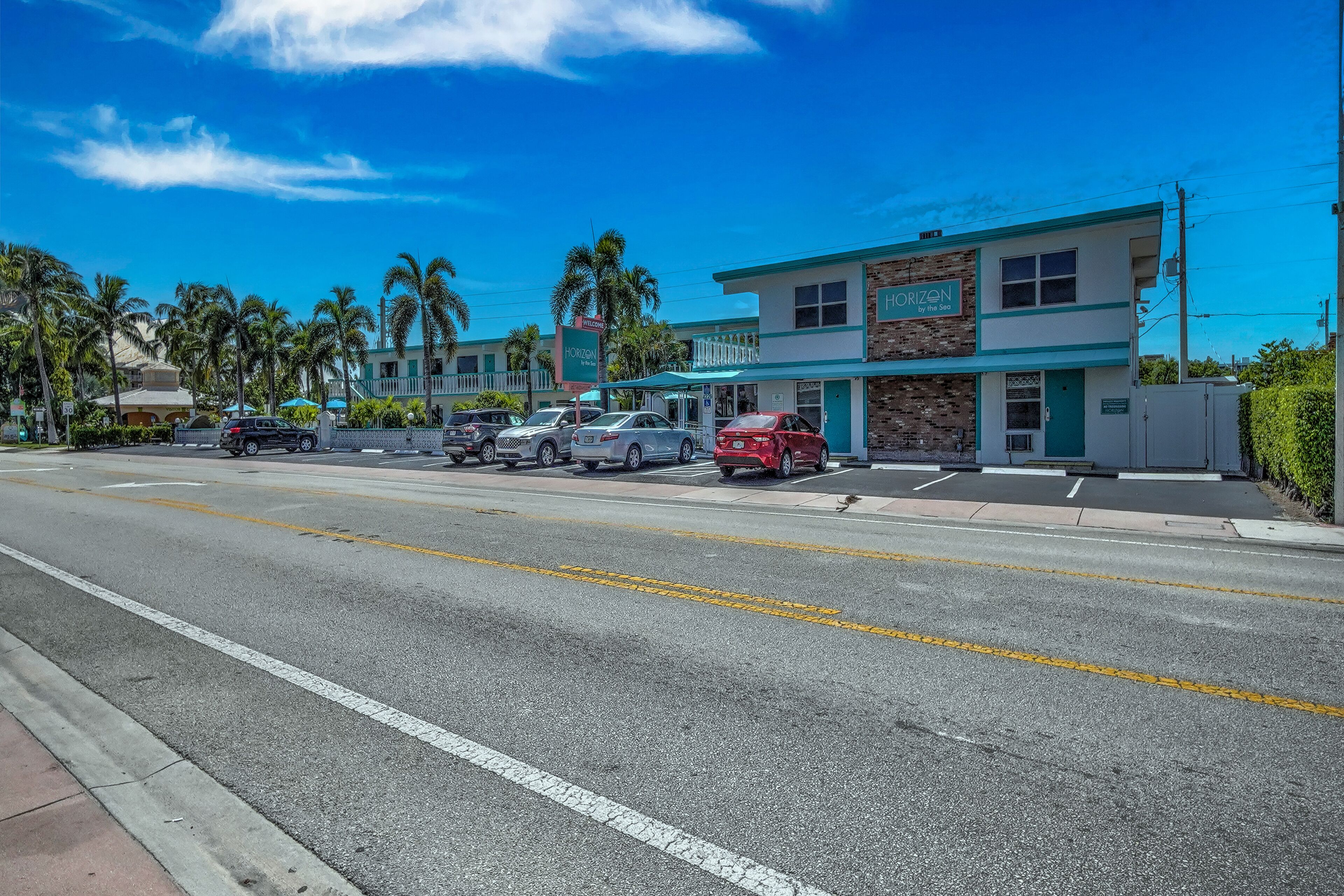 A two-story building with a sign reading "Horizon by the Sea" on the front. There are several parked cars in front of the building, and palm trees are visible in the background.