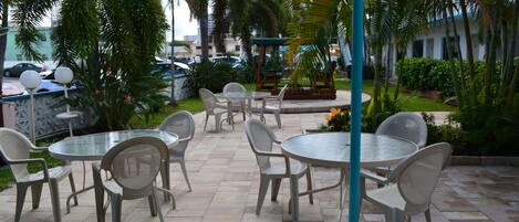 An outdoor patio with round tables and chairs, surrounded by palm trees and greenery.