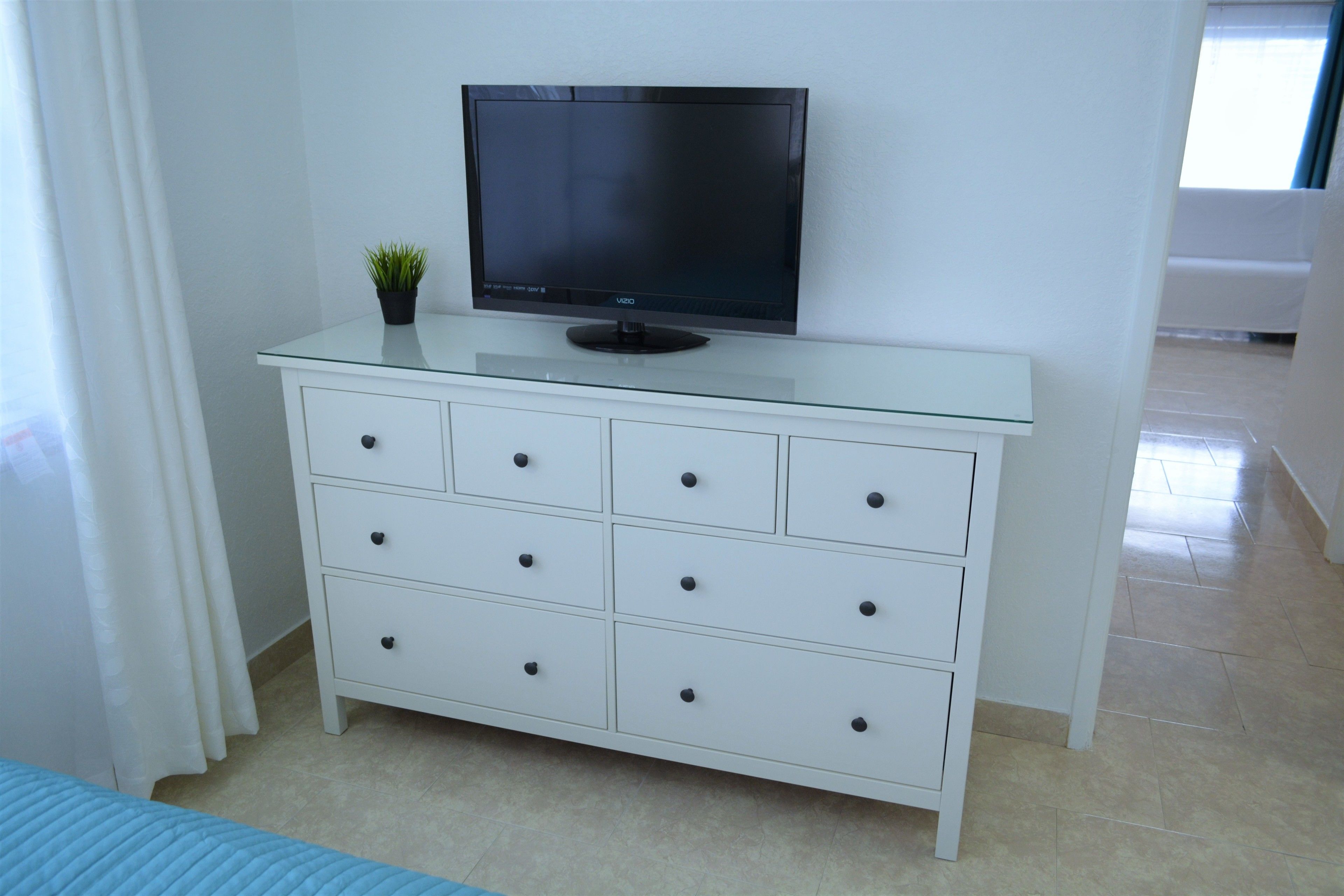 Une chambre moderne avec un téléviseur à écran plat posé sur un meuble en verre, une commode blanche dotée de nombreux tiroirs et une petite plante en pot.