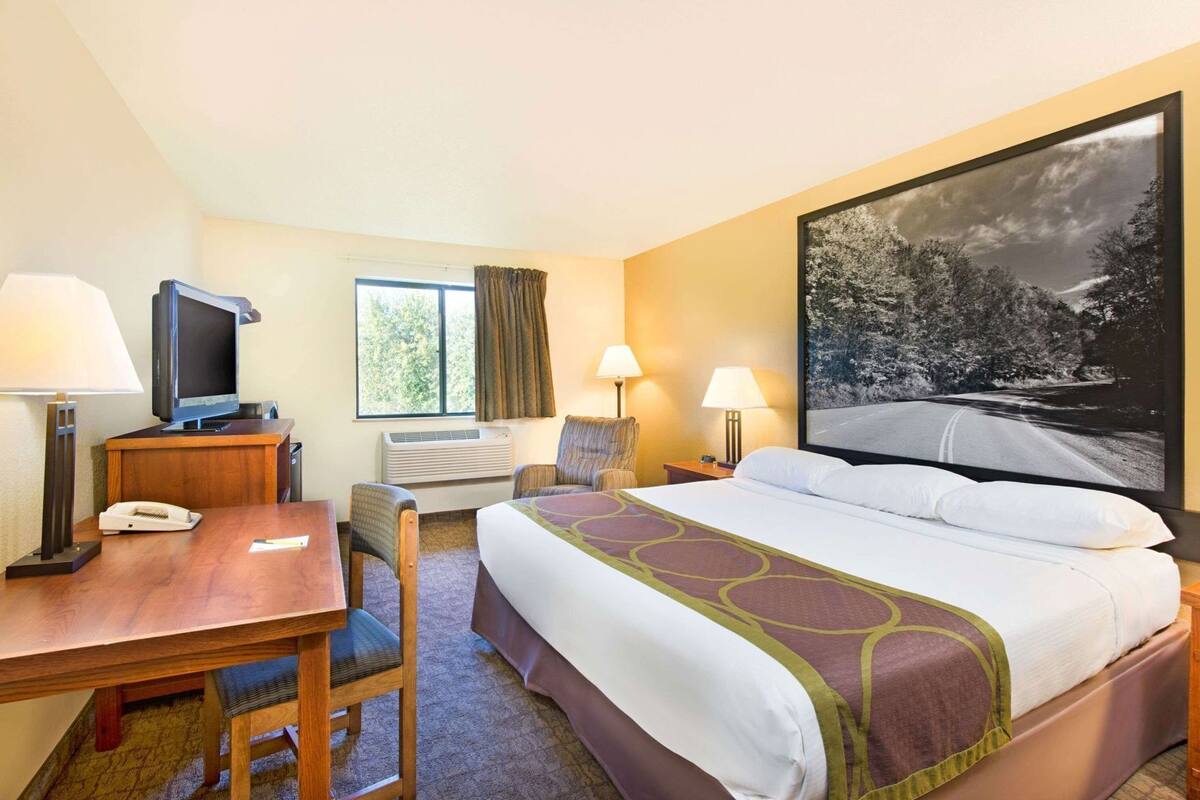 A hotel room with a bed, desk, chair, television, air conditioning unit, and a large window.