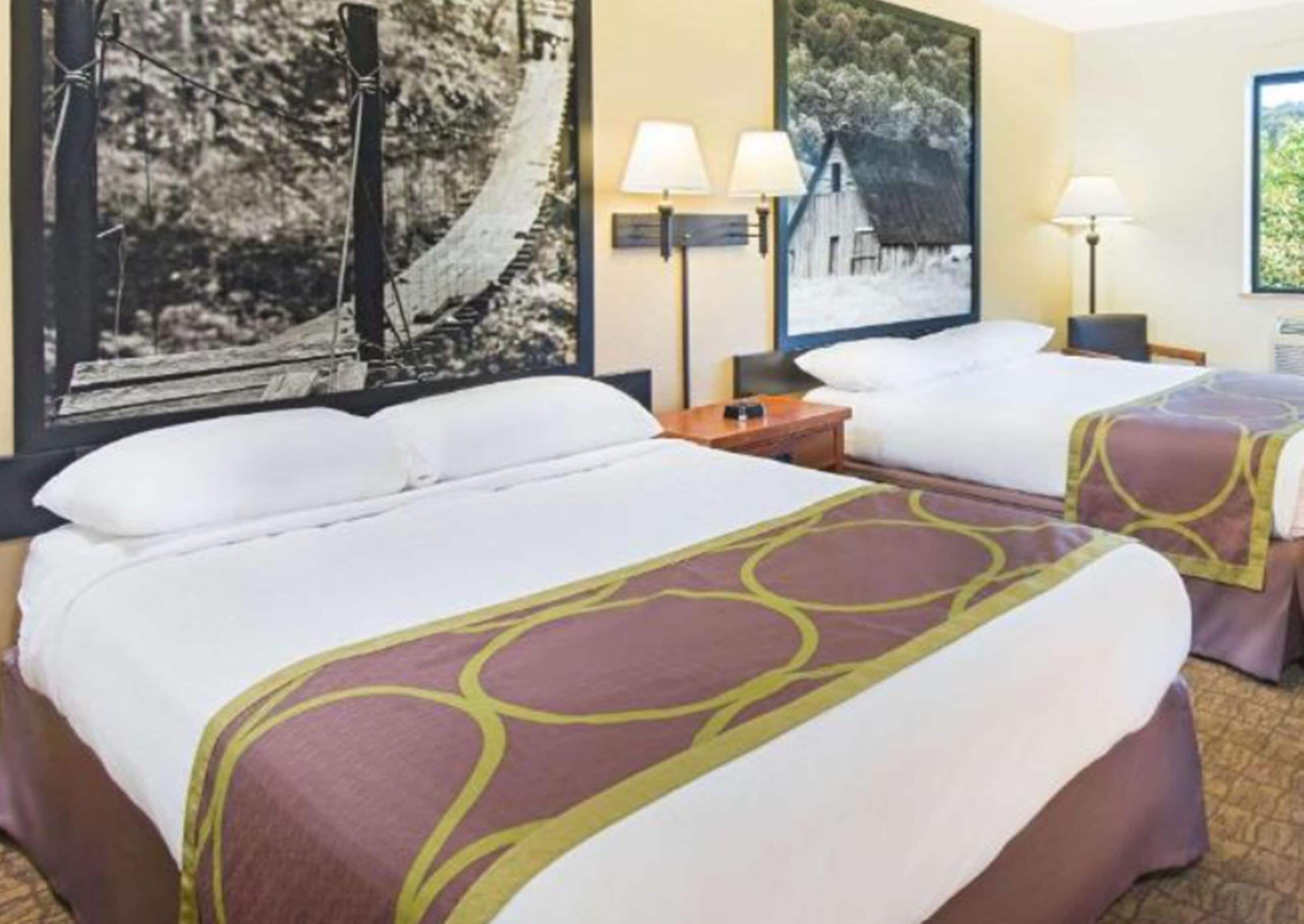 A hotel room with two beds, each with white linens and a purple and yellow patterned bedspread. There are two wall-mounted lamps on either side of the beds, and a framed picture of a rural scene hangs above them.