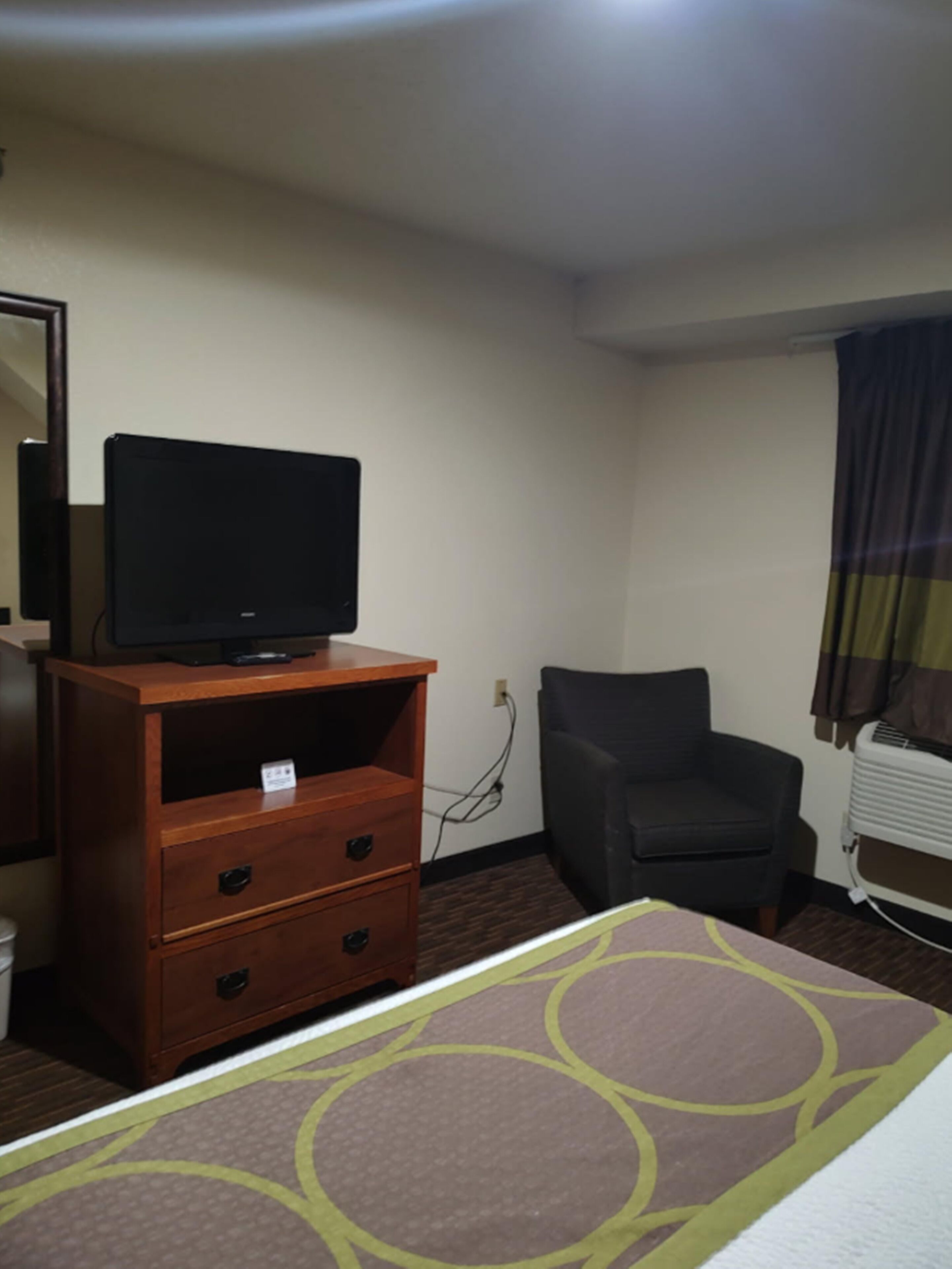 A hotel room with a wooden dresser, a television on top, a black office chair, and a patterned bedspread.