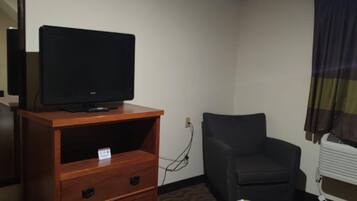 A hotel room with a wooden dresser, a television on top, a black office chair, and a patterned bedspread.