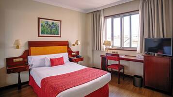 Habitación de hotel con una cama grande, un escritorio con silla, un televisor y una ventana con cortinas.