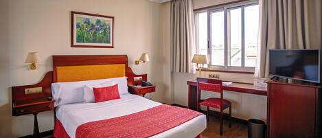 Une chambre d’hôtel avec un grand lit, un bureau avec une chaise, une télévision et une fenêtre avec des rideaux.