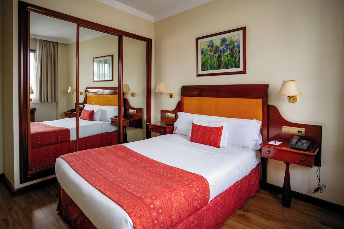 A hotel room with a large bed, a mirror, a painting, and a nightstand with a phone.