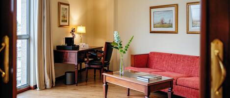 A hotel room with a wooden floor, a red sofa, a desk with a lamp, a vase with flowers, and framed pictures on the wall.