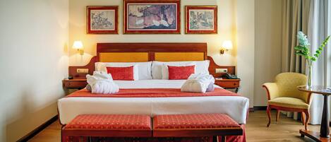 A hotel room with a large bed, two bedside tables, a chair, a small table, and three framed artworks on the wall.