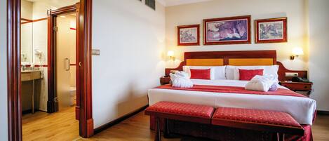 A hotel room with a wooden floor, a large bed with red bedding, a bench, and three framed artworks on the wall.