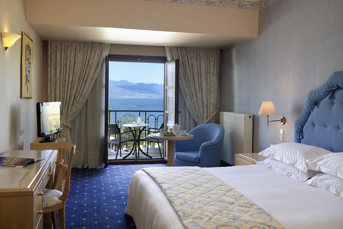 Ein Hotelzimmer mit Bett, Schreibtisch, Stuhl, Tisch mit Meerblick und einem Balkon mit Vorhängen.