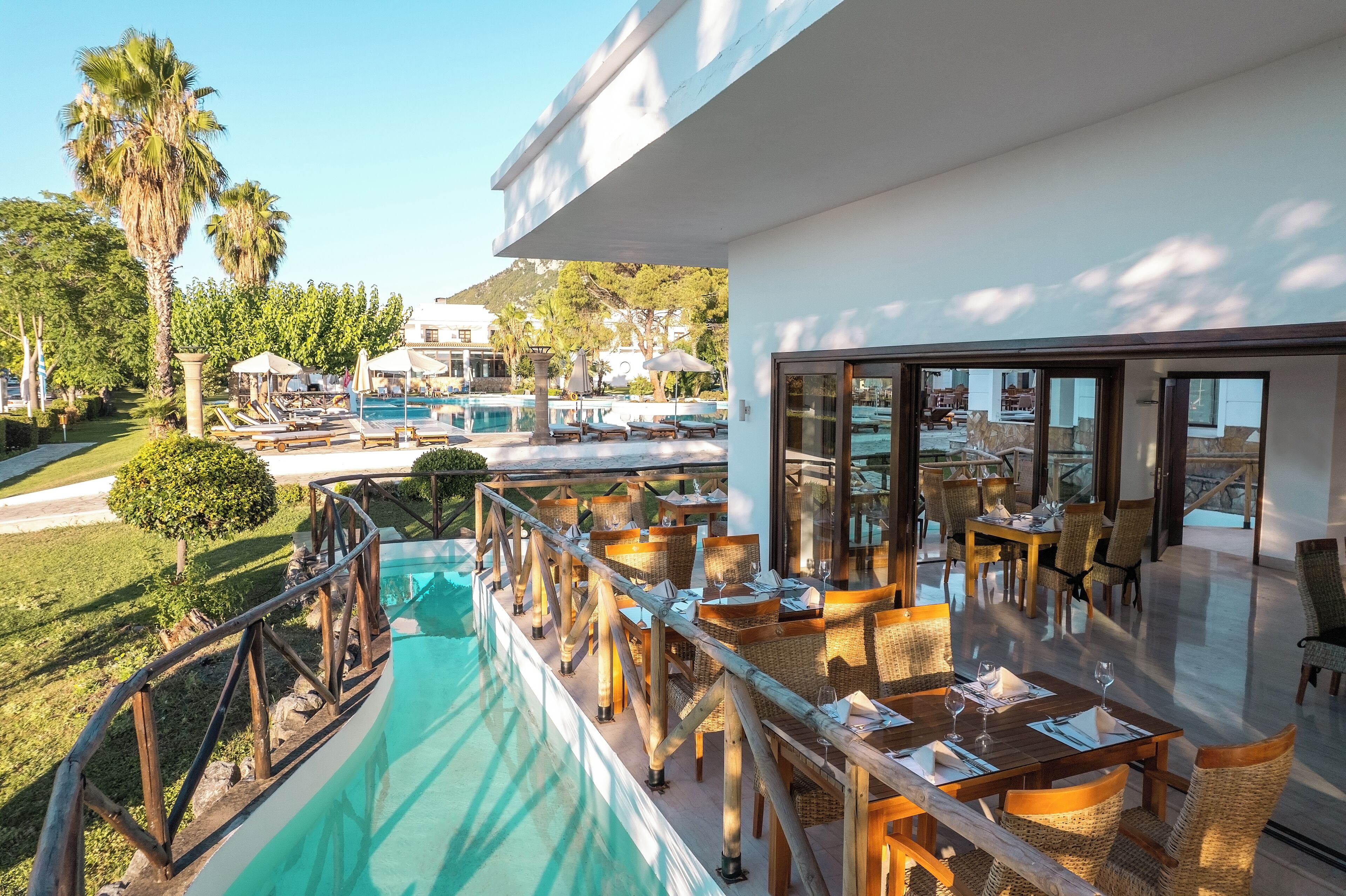 Zona junto a la piscina con sillones y mesas, vista despejada del agua y un edificio con amplias ventanas.