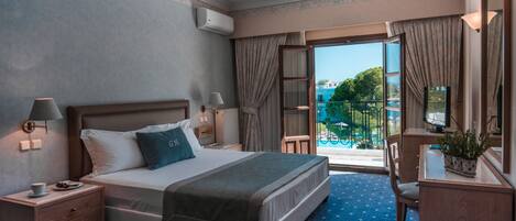 Habitación de hotel con cama, lámparas de noche, escritorio con silla, planta en maceta y vista a una piscina y vegetación.