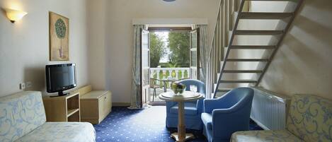 A room with a bed, a TV, a table with chairs, a balcony with a railing, and a staircase.