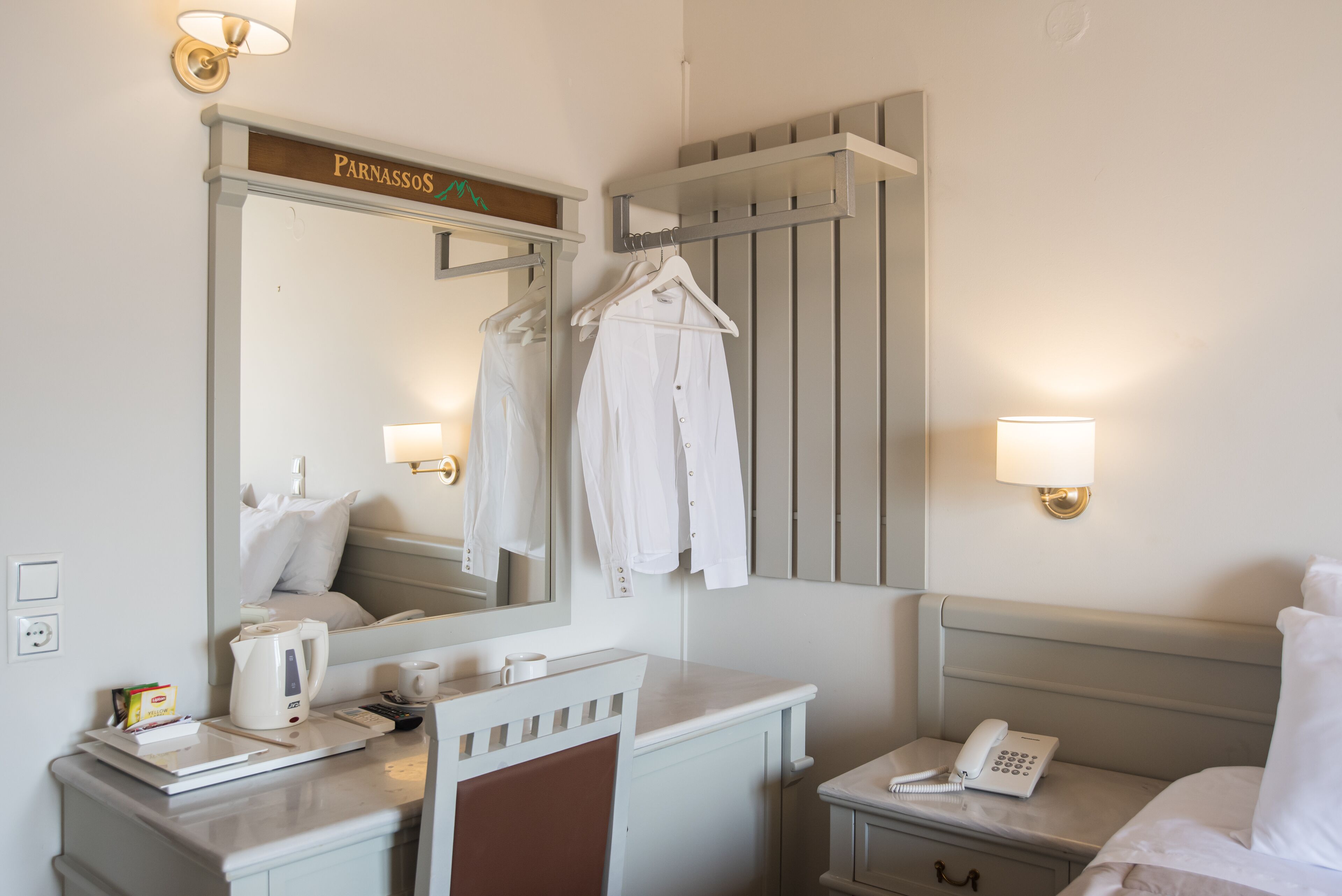 Ein Hotelzimmer mit einem Bett, einem Nachttisch mit Telefon, einem Spiegel mit dem Schild 'Pannassos', einem Handtuchhalter und einer Wandleuchte.
